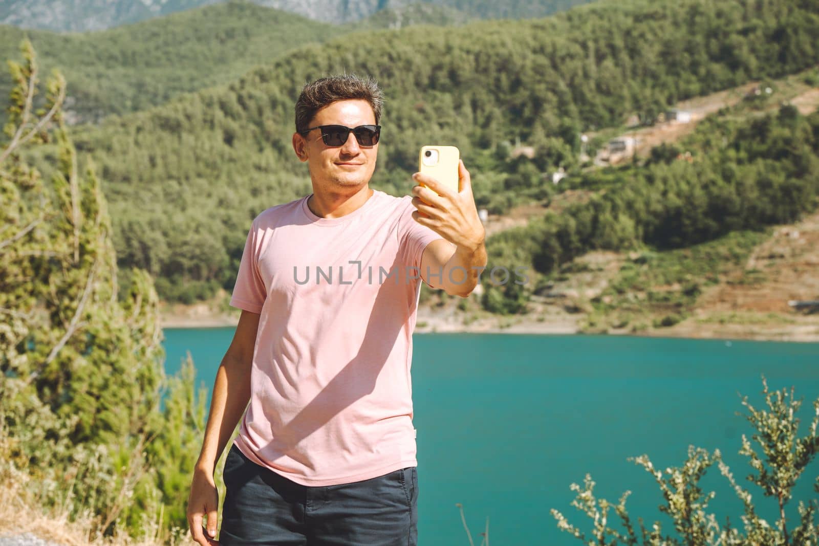 Man taking selfie on mobile phone of mountains lake background. Traveler male having video chat on cellphone on the blue lake outdoors travel adventure vacation.