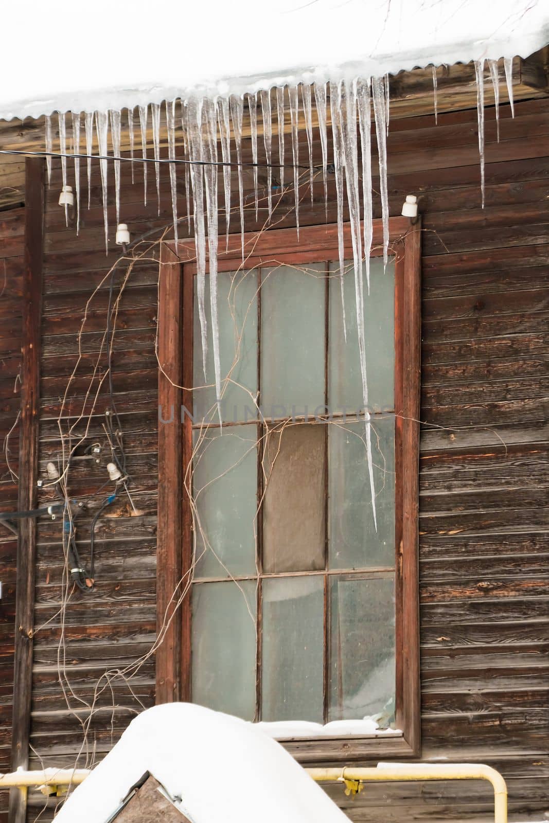 Small icicles hang on the edge of the roof, winter or spring. Log wall of an old wooden house with windows. Large cascades of icicles in smooth, beautiful rows. Cloudy winter day, soft light.