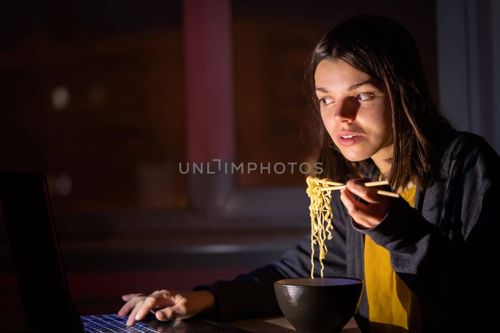 Young girl works at night at home with laptop and eats noodles, asian food with chopsticks. A woman watches a show, a movie and has dinner with ordered food from a cafe.