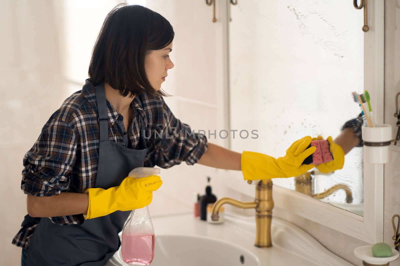 A young girl is cleaning the bathroom, applying detergent with a spray and washing the mirror with a sponge in yellow gloves on her hands. Smiling woman taking care of the cleanliness of her home.