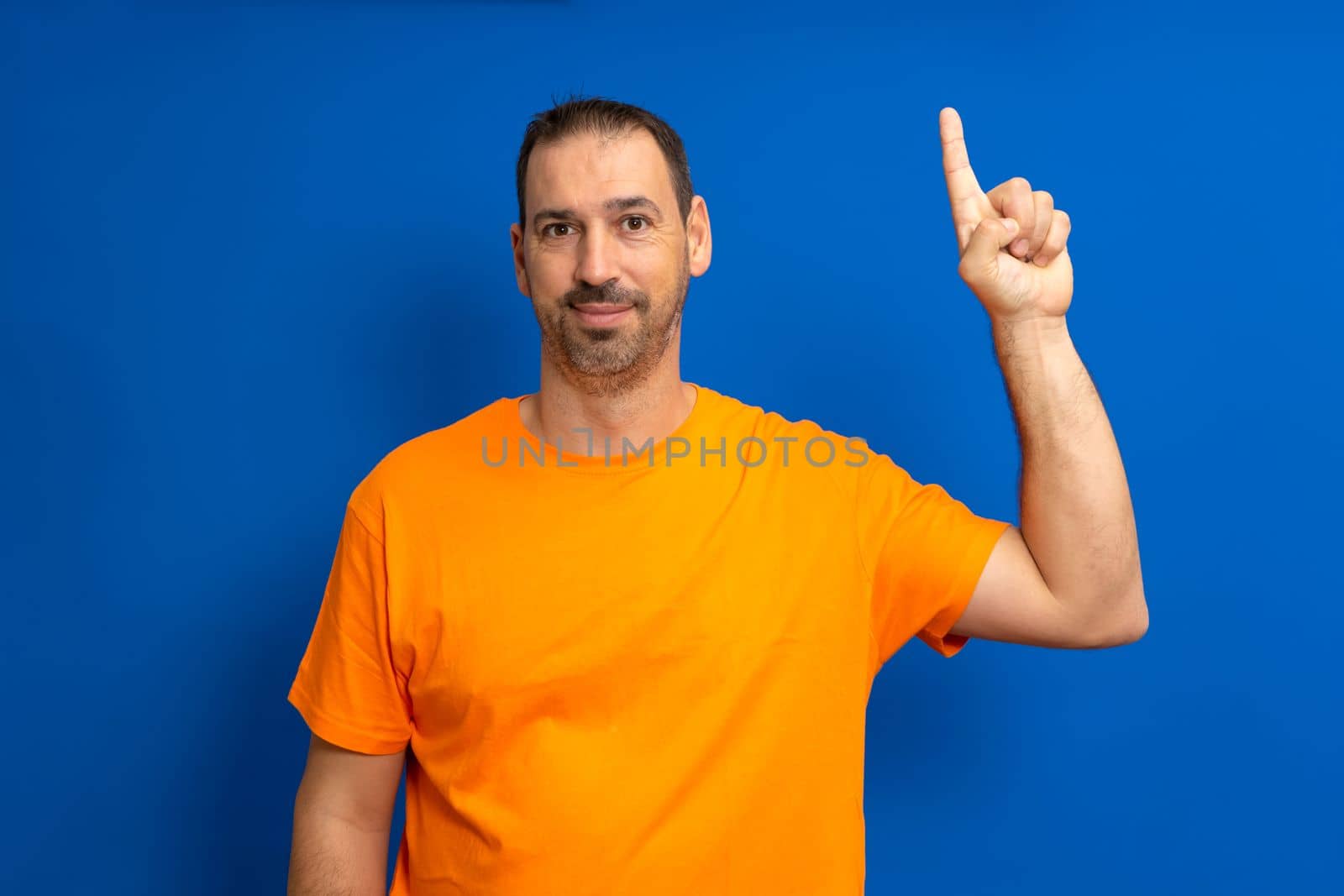 Insightful smart proactive caucasian man 40s in orange t-shirt holding index finger up with great new idea isolated on blue background studio portrait. People lifestyle concept.