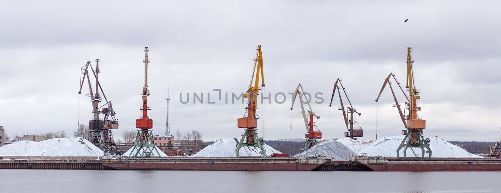 River port in winter. Port cranes on the bank of the river. Embankment of the river port, a barge at the quay wall. Large port cranes on the river. Extraction and transportation of sand other cargo