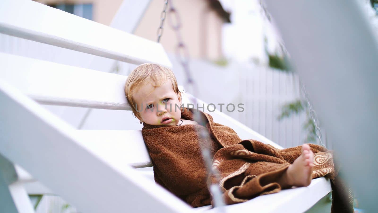 a little one-year-old girl, wrapped in a towel, with a dirty face, lonely sitting on a swing in the garden, in the summer. She has a sad look. She wants to sleep. High quality photo