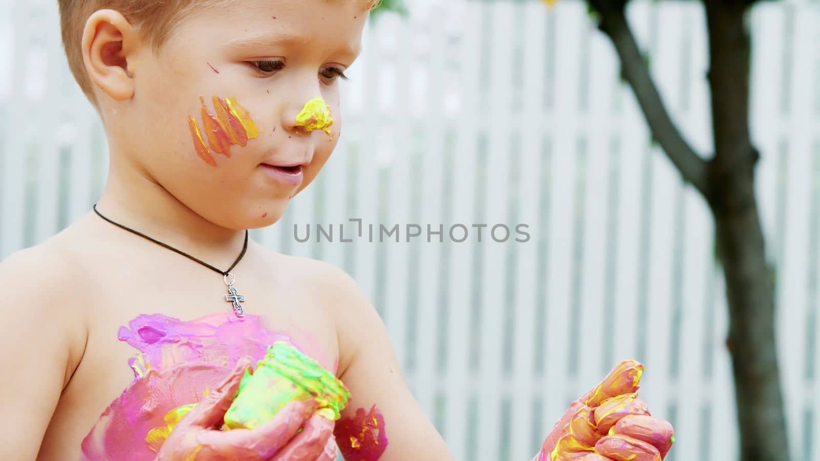 a little child, a four-year-old boy playing, painting with finger paints, decorating himself, in the garden, sitting on a blanket, on grass, lawn, in the summer. he's having fun. High quality photo