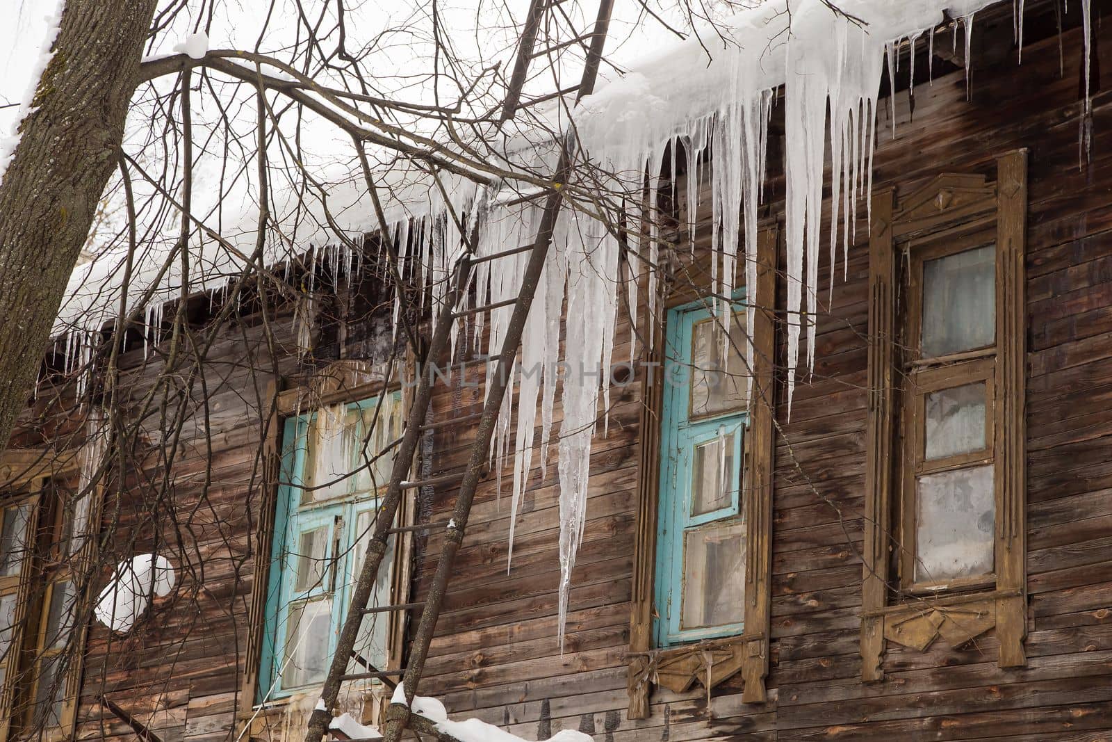 Sharp, frozen icicles hang on the edge of the roof, winter or spring. Log wall of an old wooden house with windows. Large cascades of icicles in smooth, beautiful rows. Cloudy winter day, soft light.