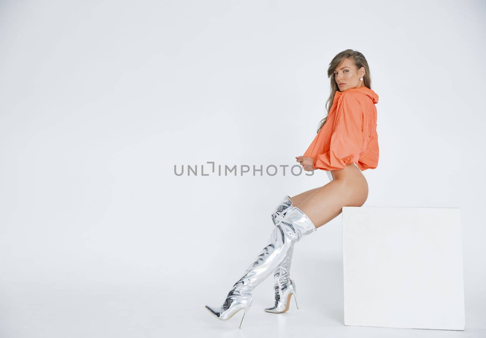 A beautiful girl of an athletic physique sits on a white cube, she is dressed in high boots of silver color, white bodysuit, pink jacket, long brown hair, white background. High quality photo