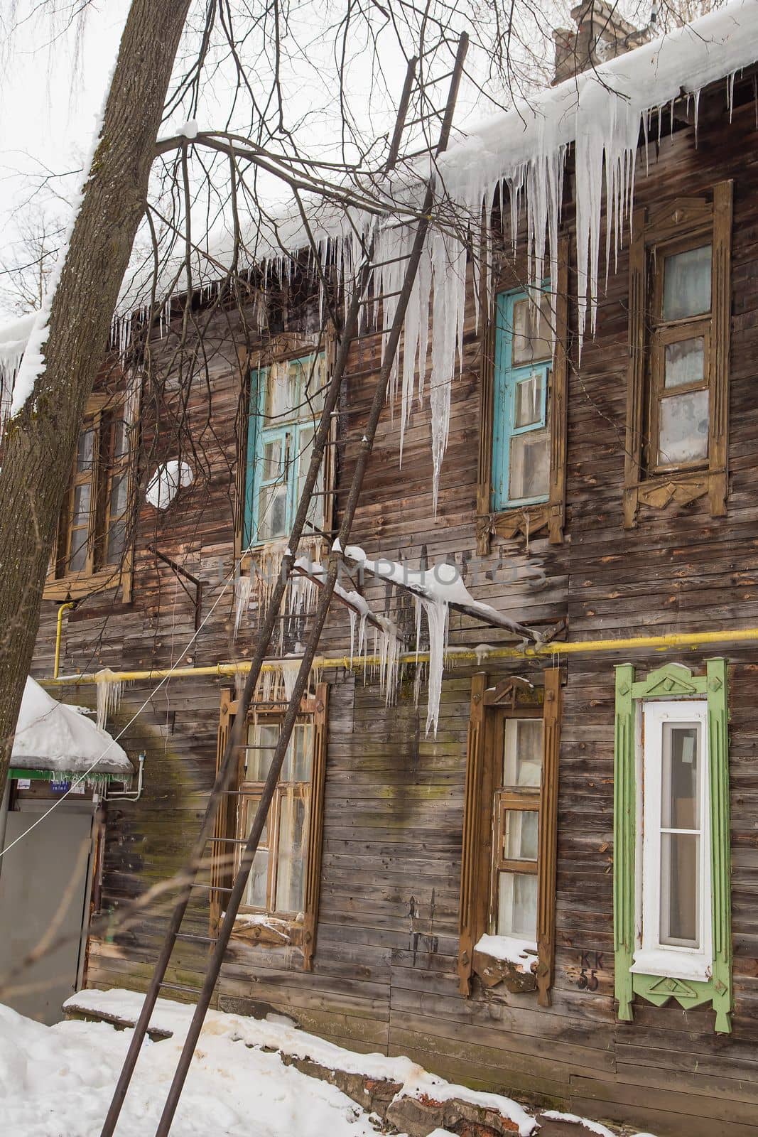 Sharp, dangerous icicles hang on the edge of the roof, winter or spring. Log wall of an old wooden house with windows. Large cascades of icicles in smooth, beautiful rows. Cloudy winter day, soft