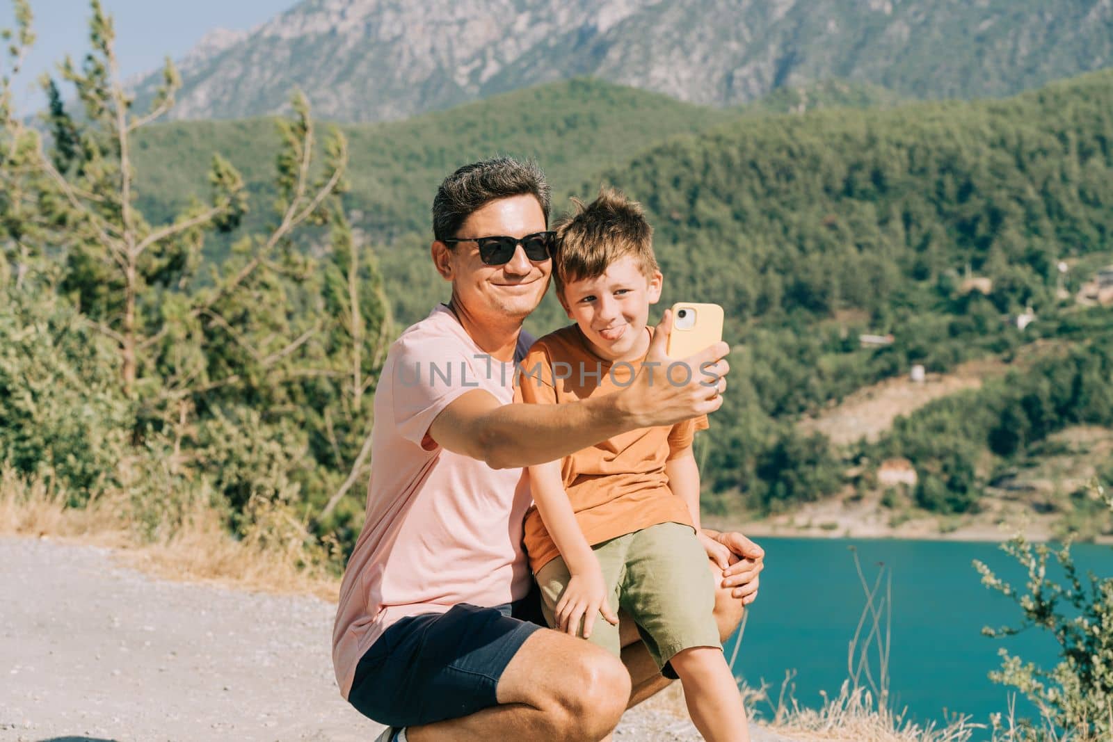 Young dad and his son standing on Mountain View and taking selfie on mobile phone. Child kid boy having fun with father and hiking hiking near mountains lake on background.