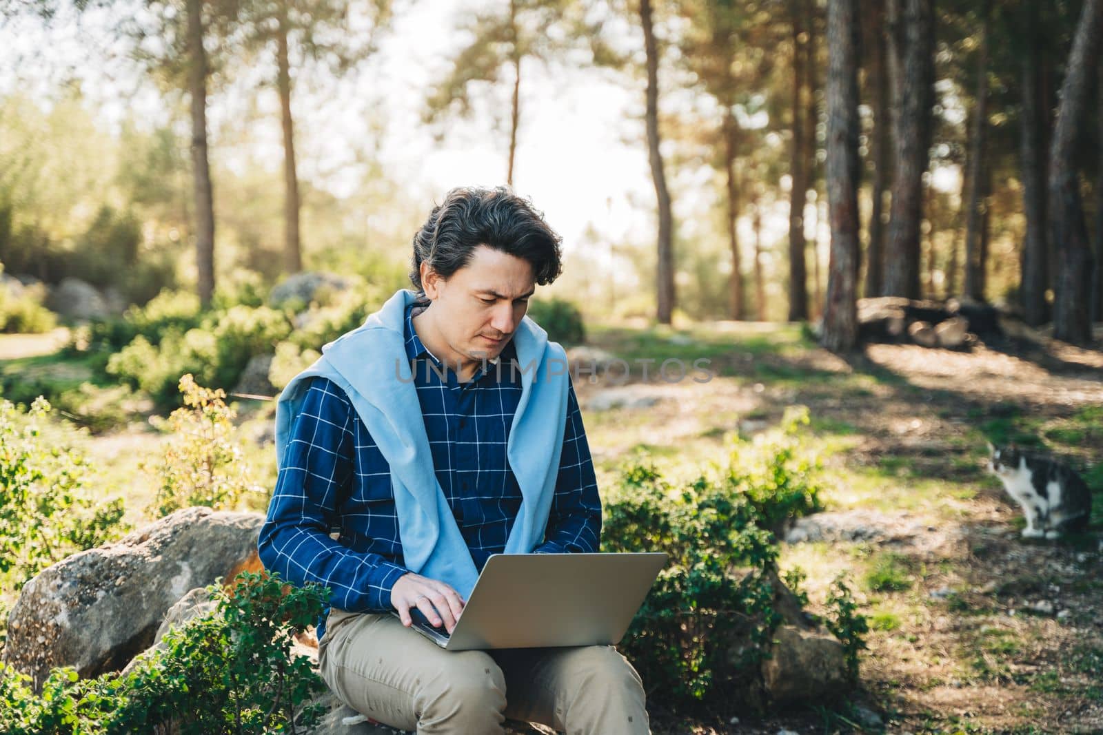 Happy cheerful man with a laptop sitting outdoors on rock in nature forest park. Freelancer with computer typing, blogging, browsing in vacation in mountains. Freelance, distance work concept by Ostanina