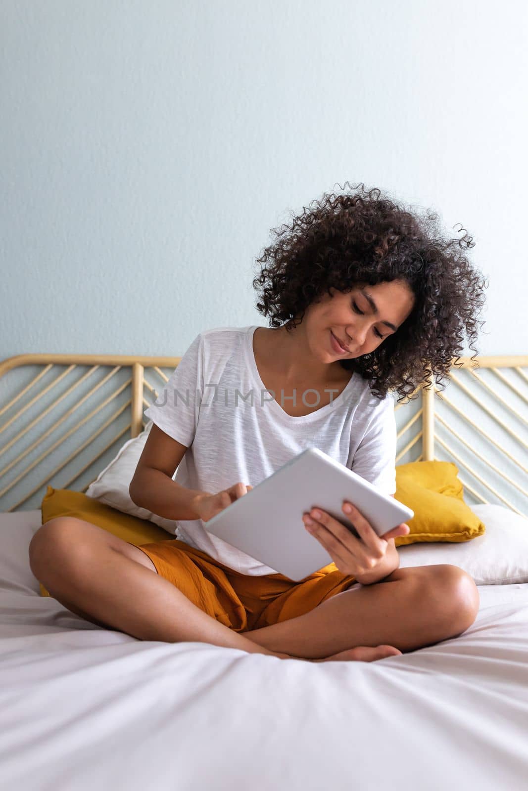 Young multiracial woman using sitting on bed down relaxing at home cozy bedroom. Vertical image.Technology concept.