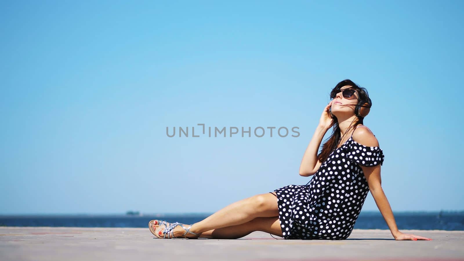 beautiful brunette girl in sun glasses and sundress, with big headphones, listening to music from smartphone, on beach, on hot summer day, near sea. High quality photo