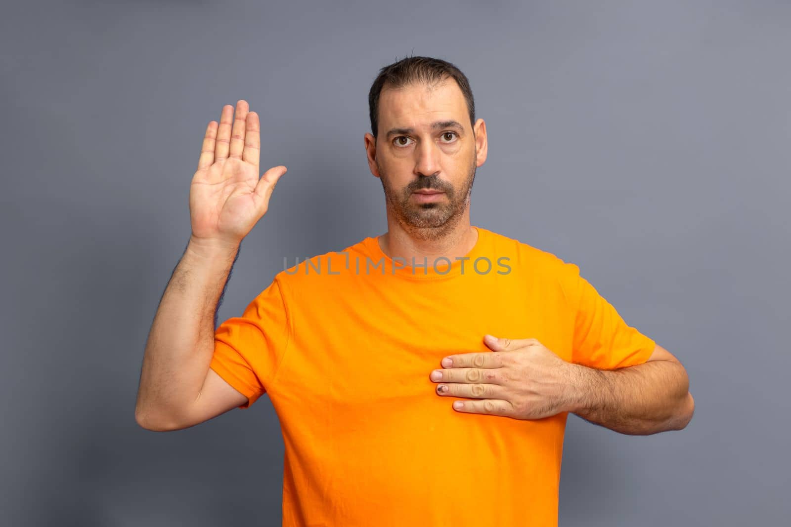 Bearded hispanic man wearing orange t-shirt isolated on gray background swearing, putting hand on chest.