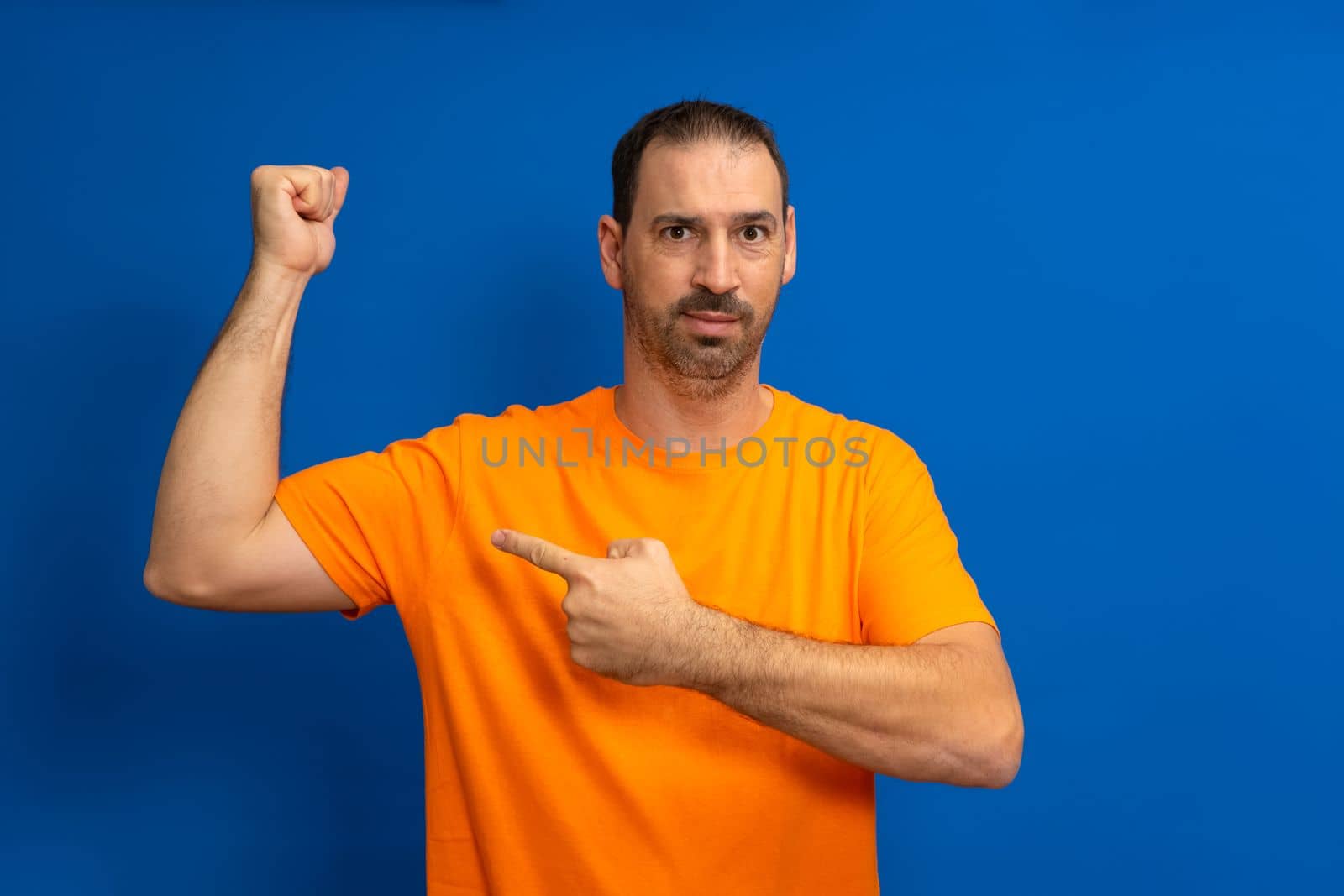 Handsome funny bearded latino man pointing at his biceps and smiling over blue background.