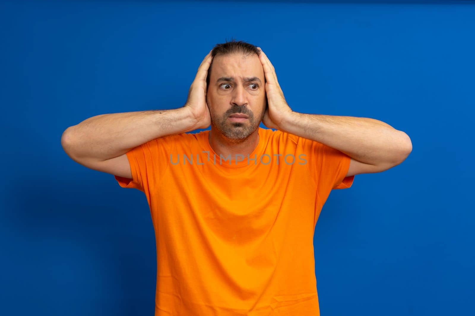 Stunned latin man wearing an orange t-shirt with his head in his hands isolated on a blue studio background