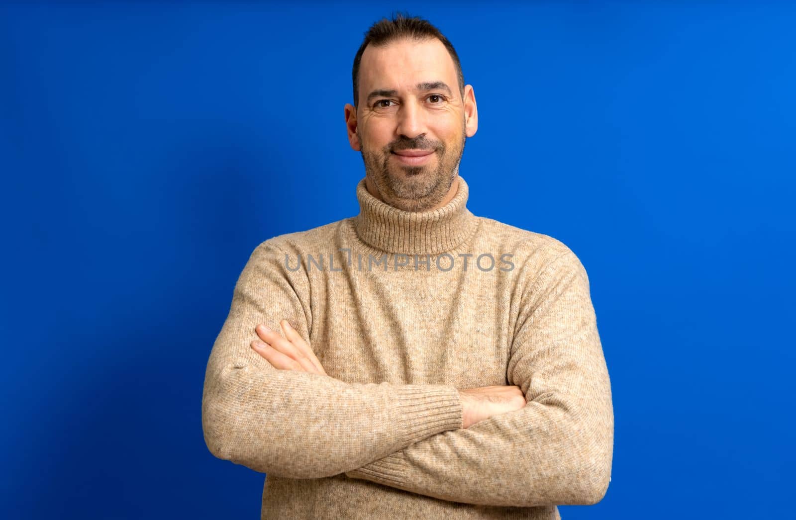 Smiling handsome man in a brown turtleneck standing with arms crossed isolated on blue studio background