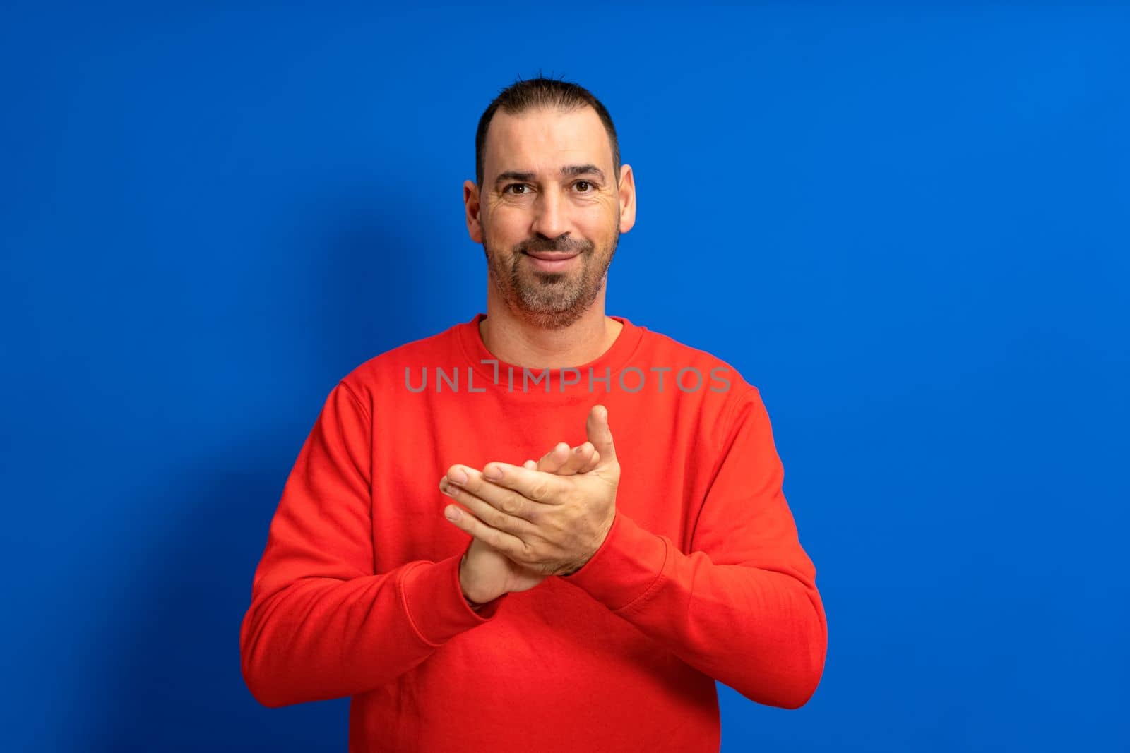Handsome hispanic man with a beard in a red jumper clapping his hands in comfort after witnessing a nice show isolated over blue studio background