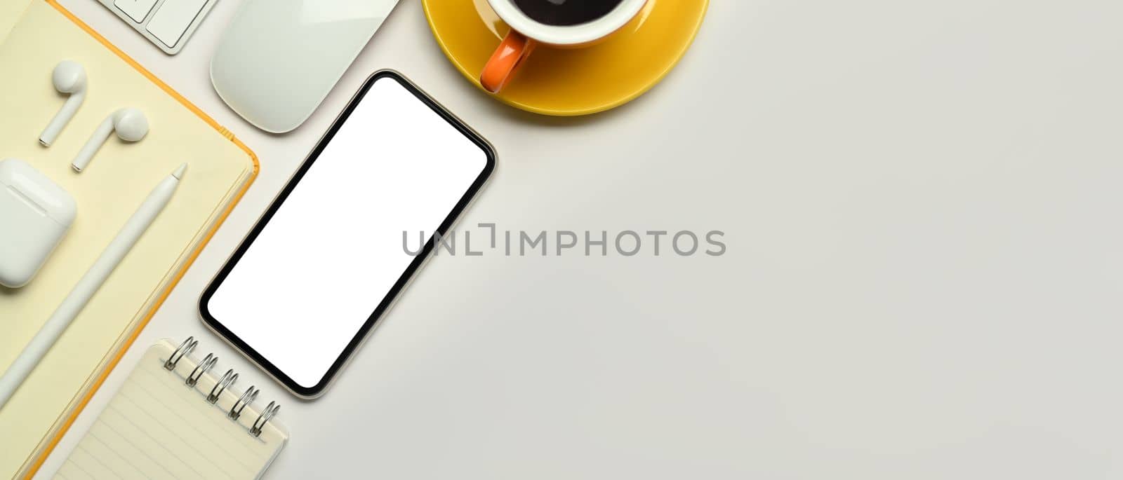 Mock up smart phone with empty screen, notepad, coffee cup and wireless earphones on white table.