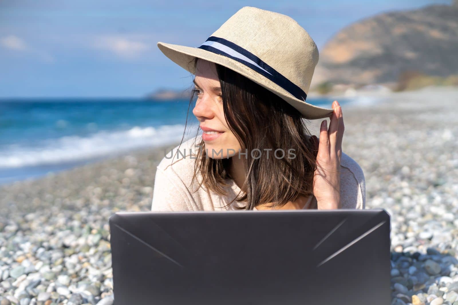 A young girl in a light hat and casual sweater lies on the beach by the sea with a laptop on a sunny day, works, studies, buys tickets during trip, a woman rests on vacation and types on the keyboard.