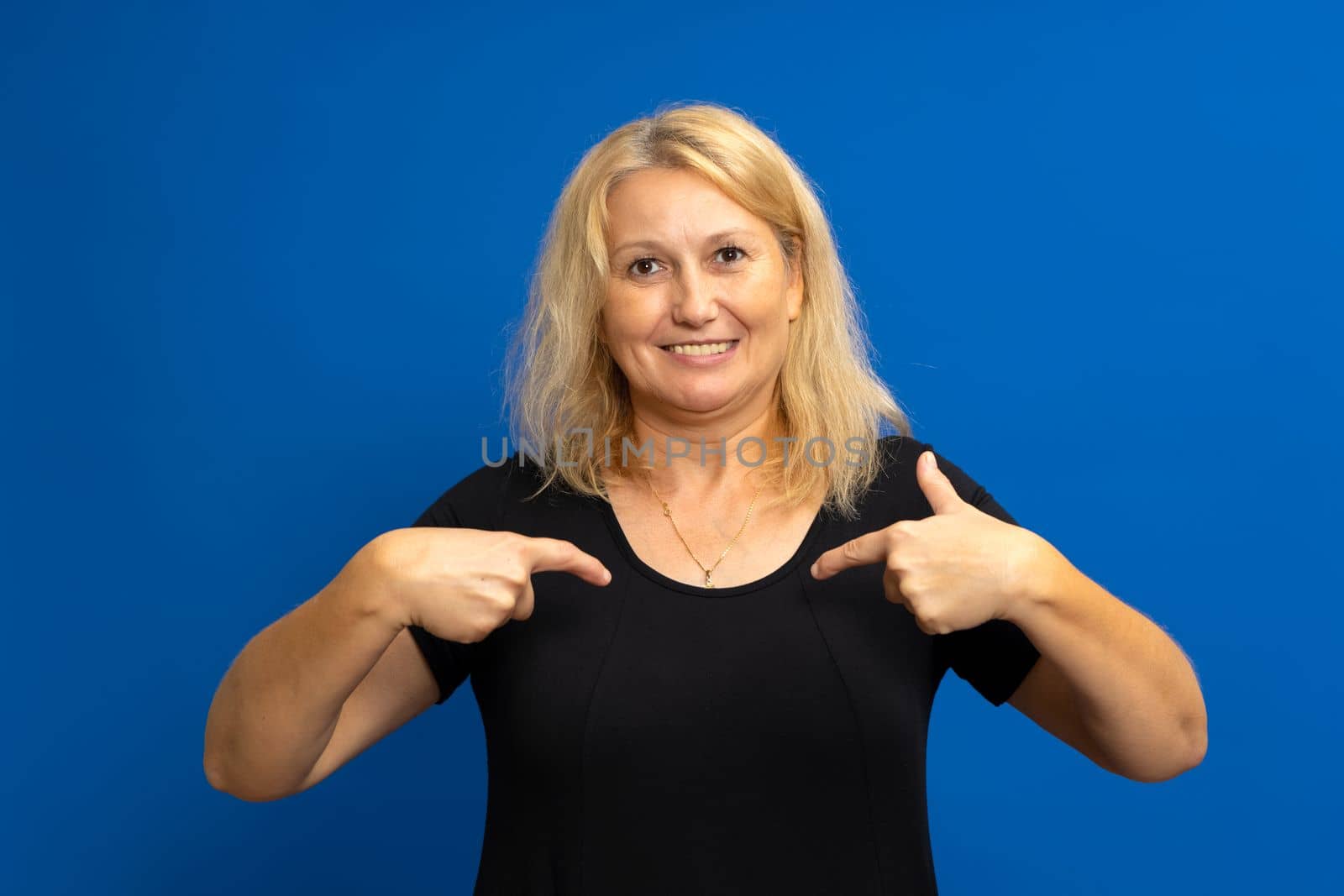 Portrait of a charming happy woman in a black t-shirt smiling and pointing at herself, proud of her own success, bragging about her achievements. Indoor studio shot isolated on blue background.