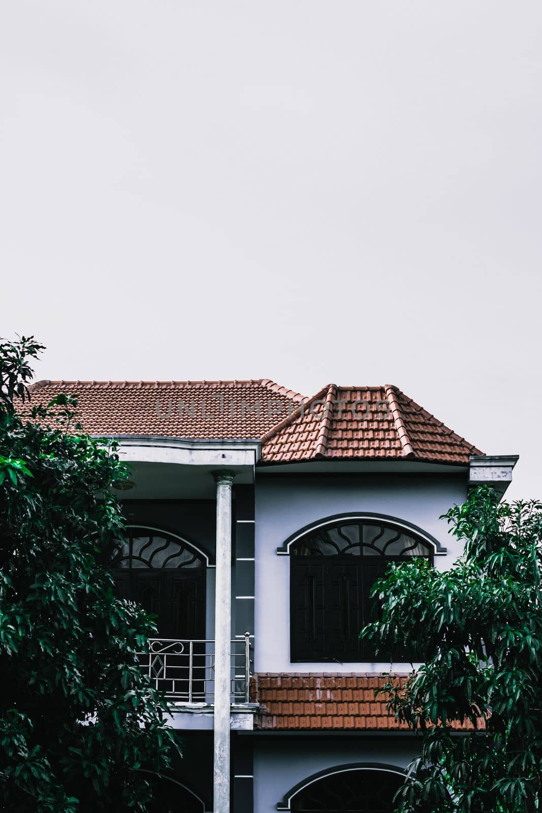 Vertical photo modern roof red tiles two-story building top floor trees green light clear background sky.