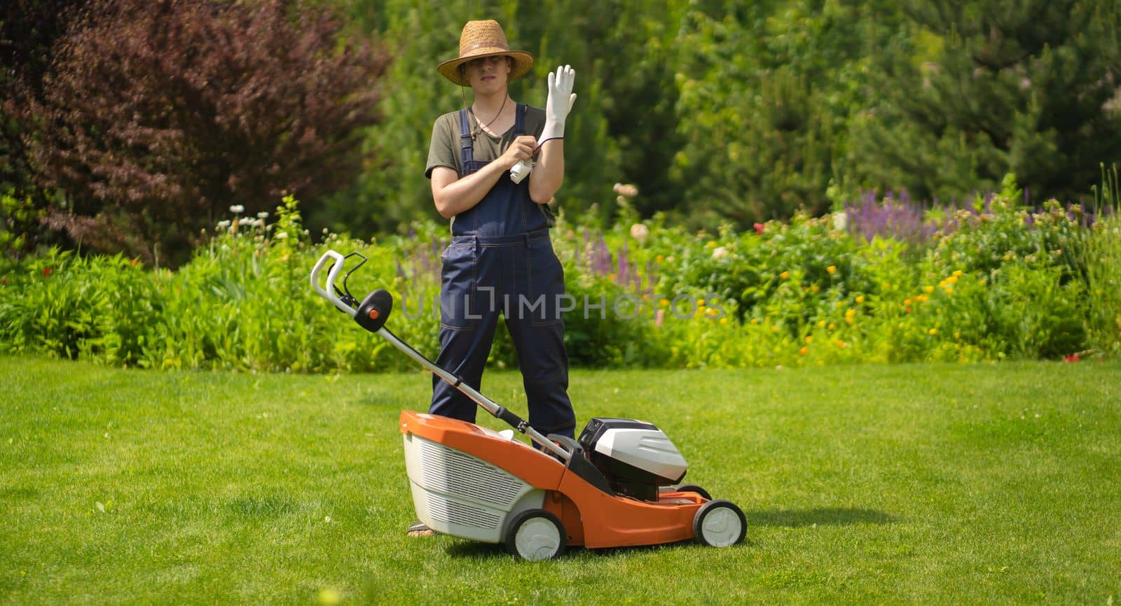 A young man in a straw hat is putting on gloves to mowe a lawn with a lawn mower in his beautiful green floral summer garden. A professional gardener with a lawnmower cares for the grass in the backyard.