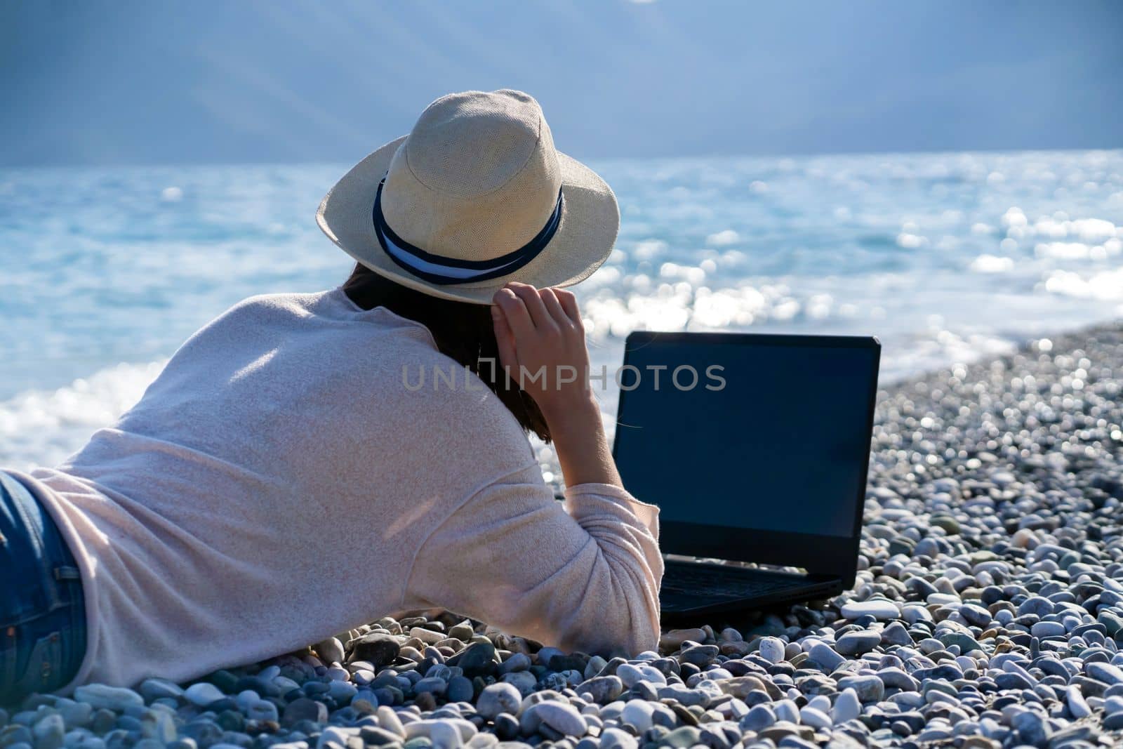 A young girl in a light hat and casual sweater lies on the beach by the sea with a laptop on a sunny day, works, studies, buys tickets during trip, a woman rests on vacation and types on the keyboard.