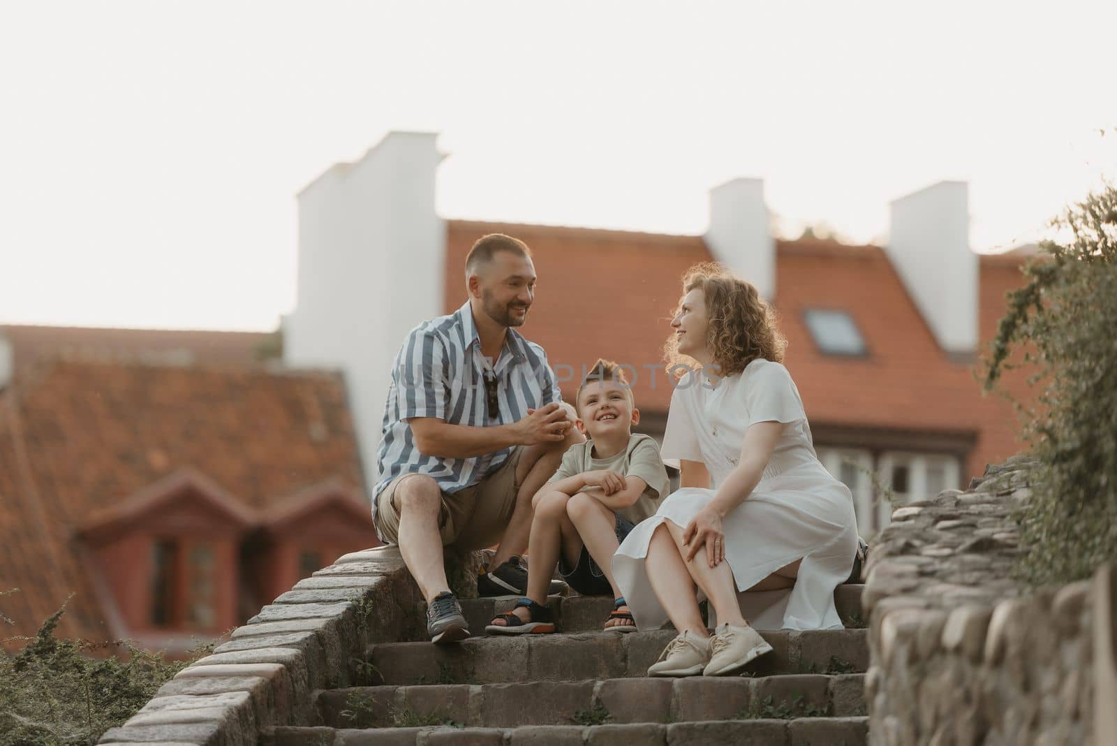 A smiling family is joking on the stairs between roofs in an old European town. A happy father, mother, and son are having fun in the evening.