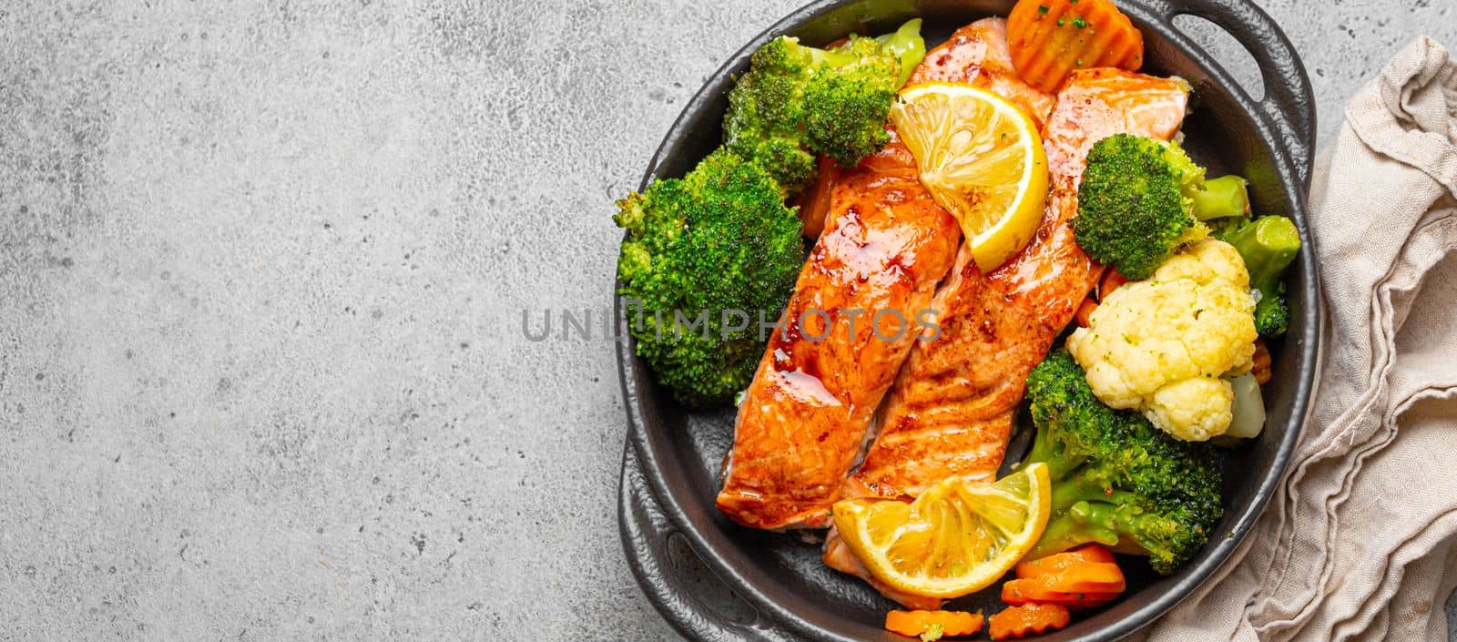 Healthy baked fish salmon steaks, broccoli, cauliflower, carrot in black cast iron casserole bowl on grey stone background. Cooking a delicious low carb dinner, healthy nutrition, space for text