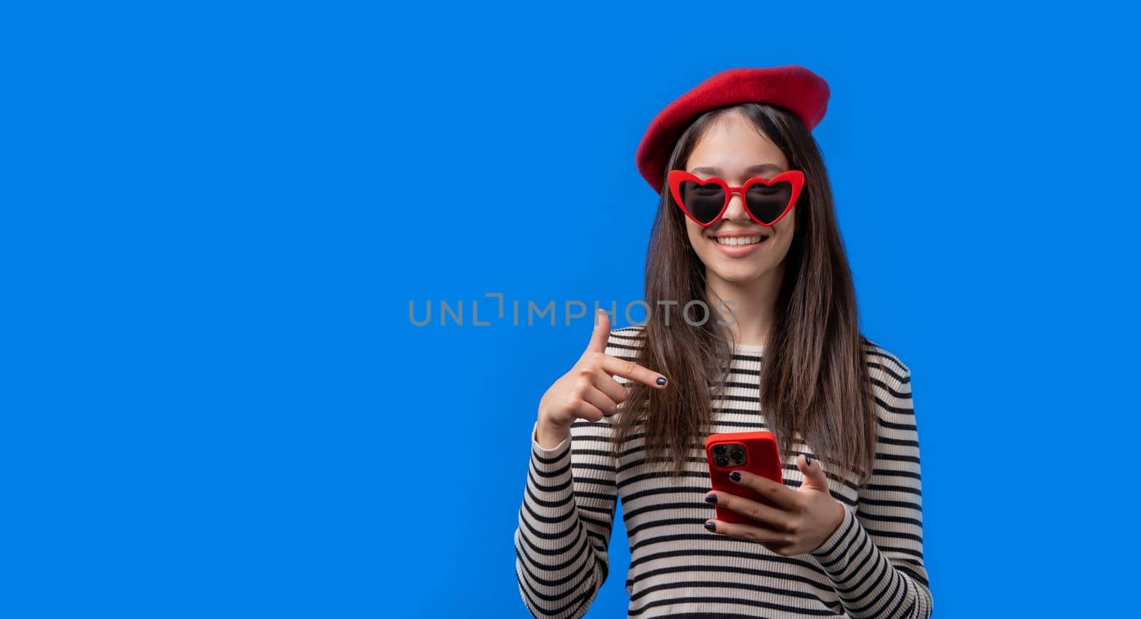 Smiling teenager girl pointing to smartphone, she advise mobile app, mobile operator. Young woman on blue background. Banner, copy space. High quality photo