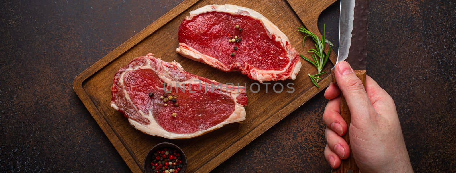 Male's hand holding knife over two raw uncooked meat beef rib eye marbled steaks on wooden cutting board and seasonings on dark rustic background from above, preparing dinner with meat