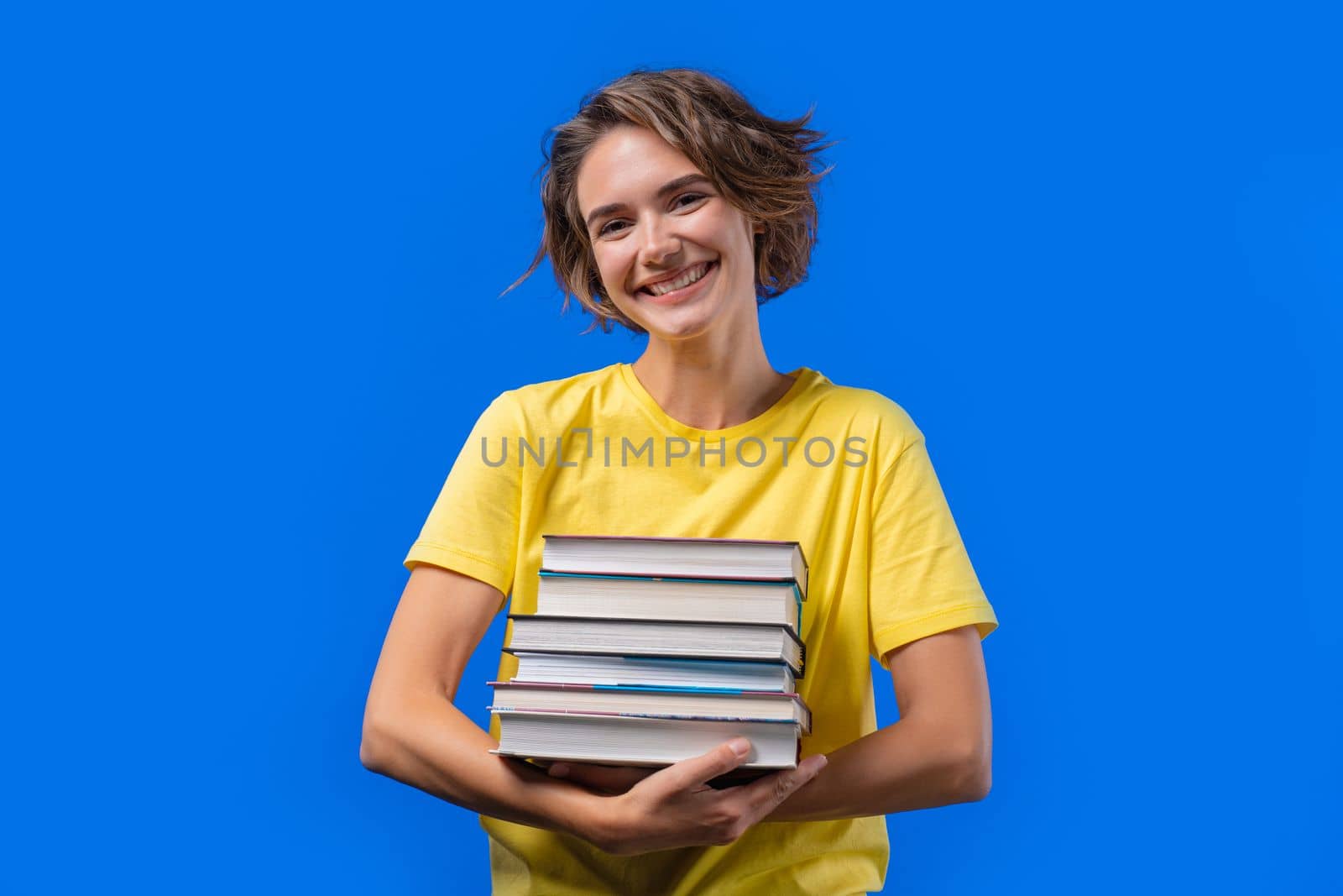 Clever student woman holds stack of university books from college library on blue background. Happy girl smiles, she is happy to graduate. by kristina_kokhanova