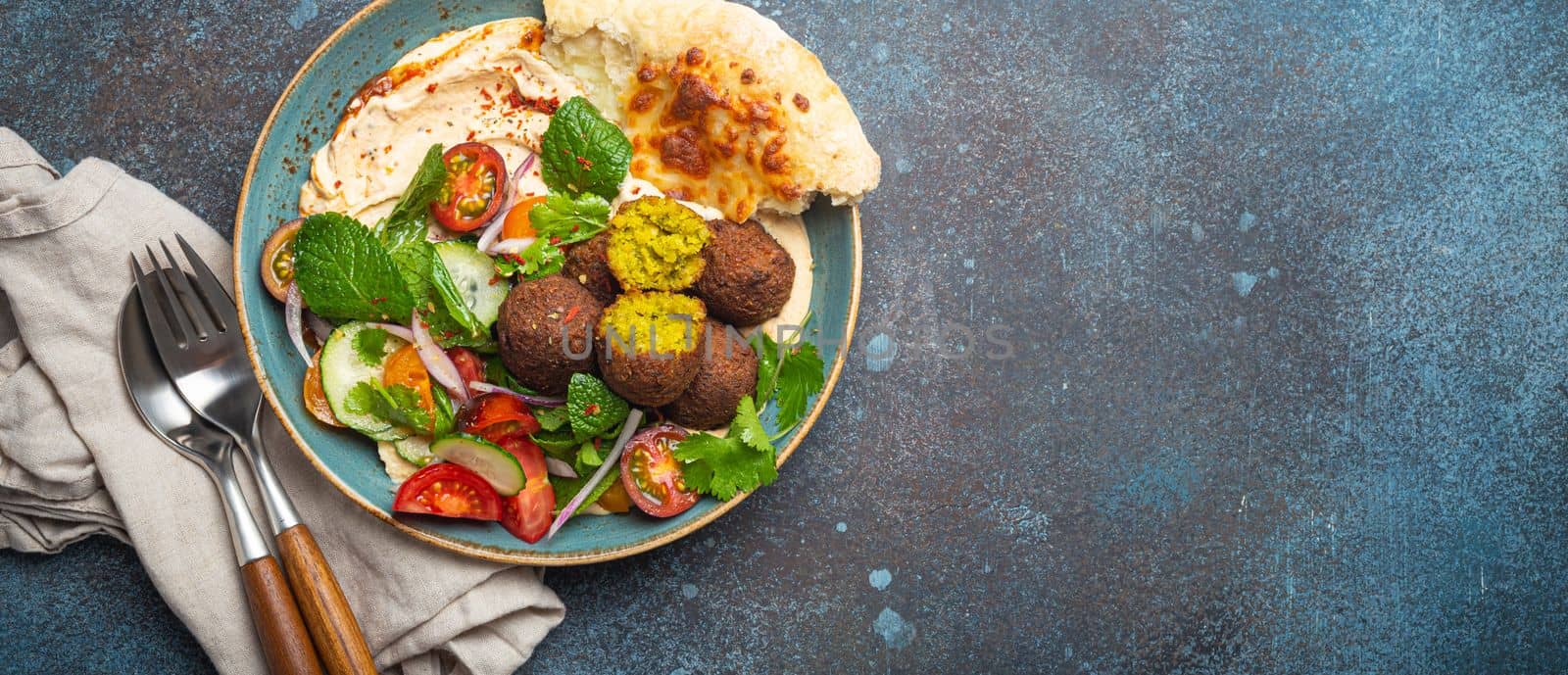 Middle Eastern Arab meal with fried falafel, hummus, vegetables salad with fresh green cilantro and mint leaves, pita bread in ceramic bowl on stone rustic background top view, space for text