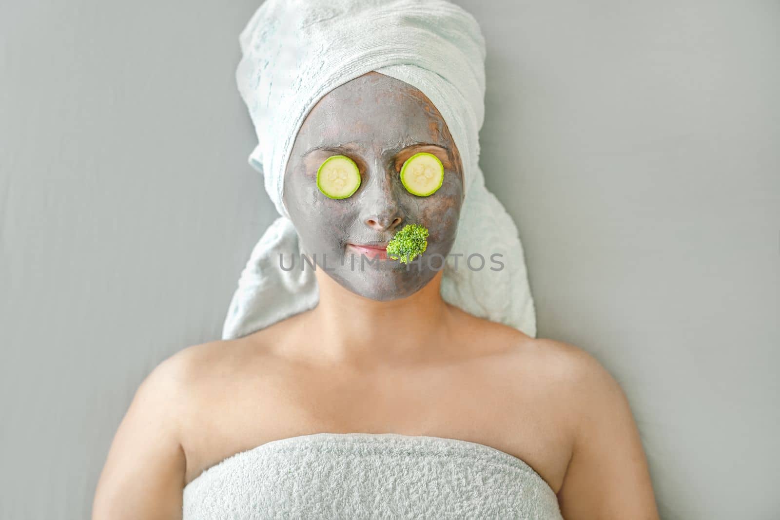 Spa procedure. Funny girl with cosmetic mask made of gray clay and pieces of vegetables on her face, her hair wrapped in towels. Concept of natural facial skin care. by Laguna781