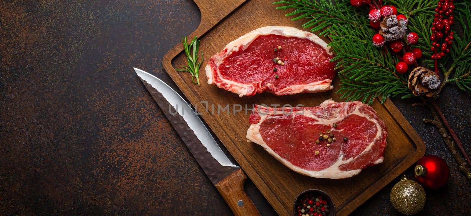 Two raw uncooked meat beef steaks on wooden cutting board with knife and seasonings on dark rustic background with Christmas festive decoration from above with space for text