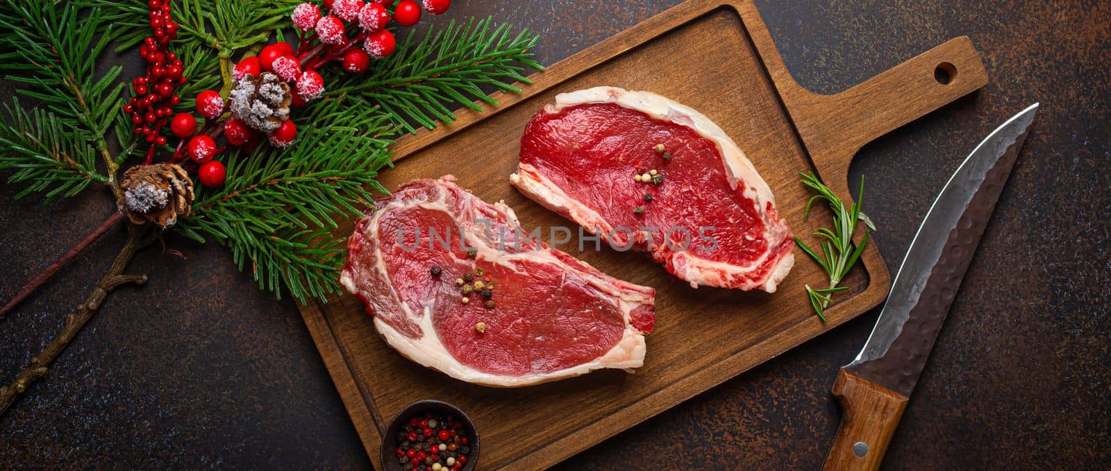 Two raw uncooked meat beef steaks on wooden cutting board with knife and seasonings on dark rustic background with Christmas festive decoration from above