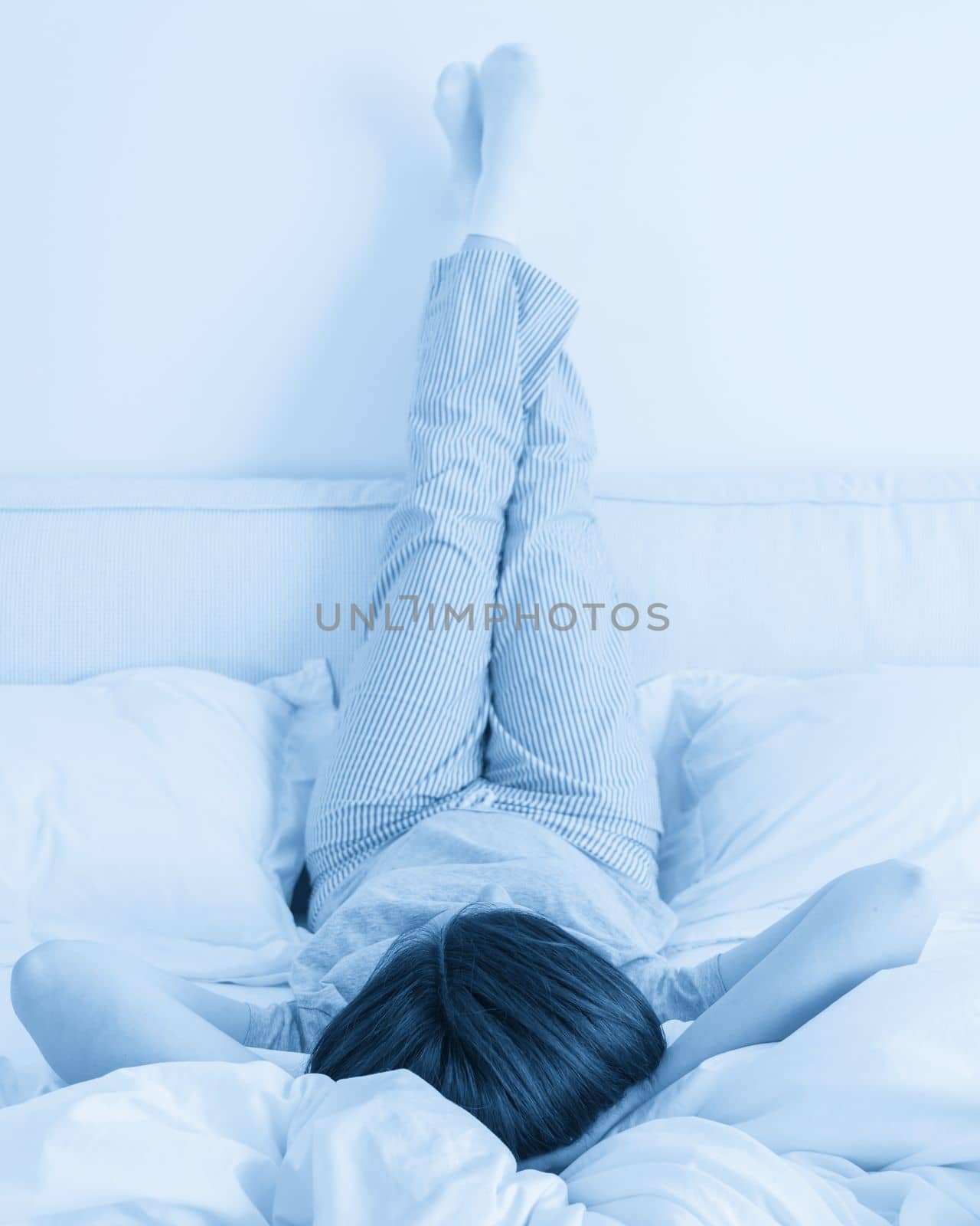 Female legs raised up high and arms under her head lying on bed in bedroom wearing pajamas by Mariakray