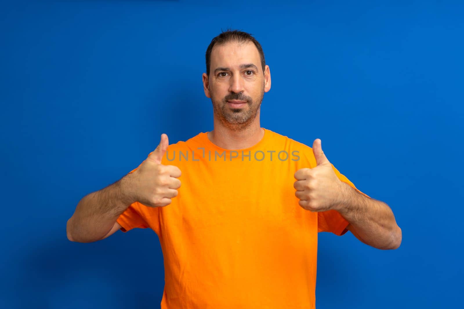 Portrait of a cheerful bearded latino man in orange t-shirt smiling and showing thumbs up to camera isolated over blue background
