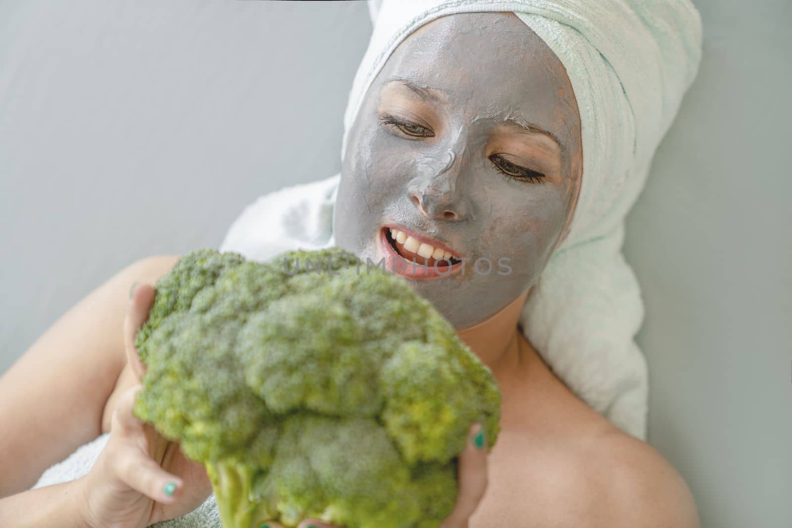 Funny girl with cosmetic mask made of gray clay, her hair and body wrapped in towels, she holds broccoli in her hands, portrait, close-up. Spa procedure, concept of natural facial skin care.