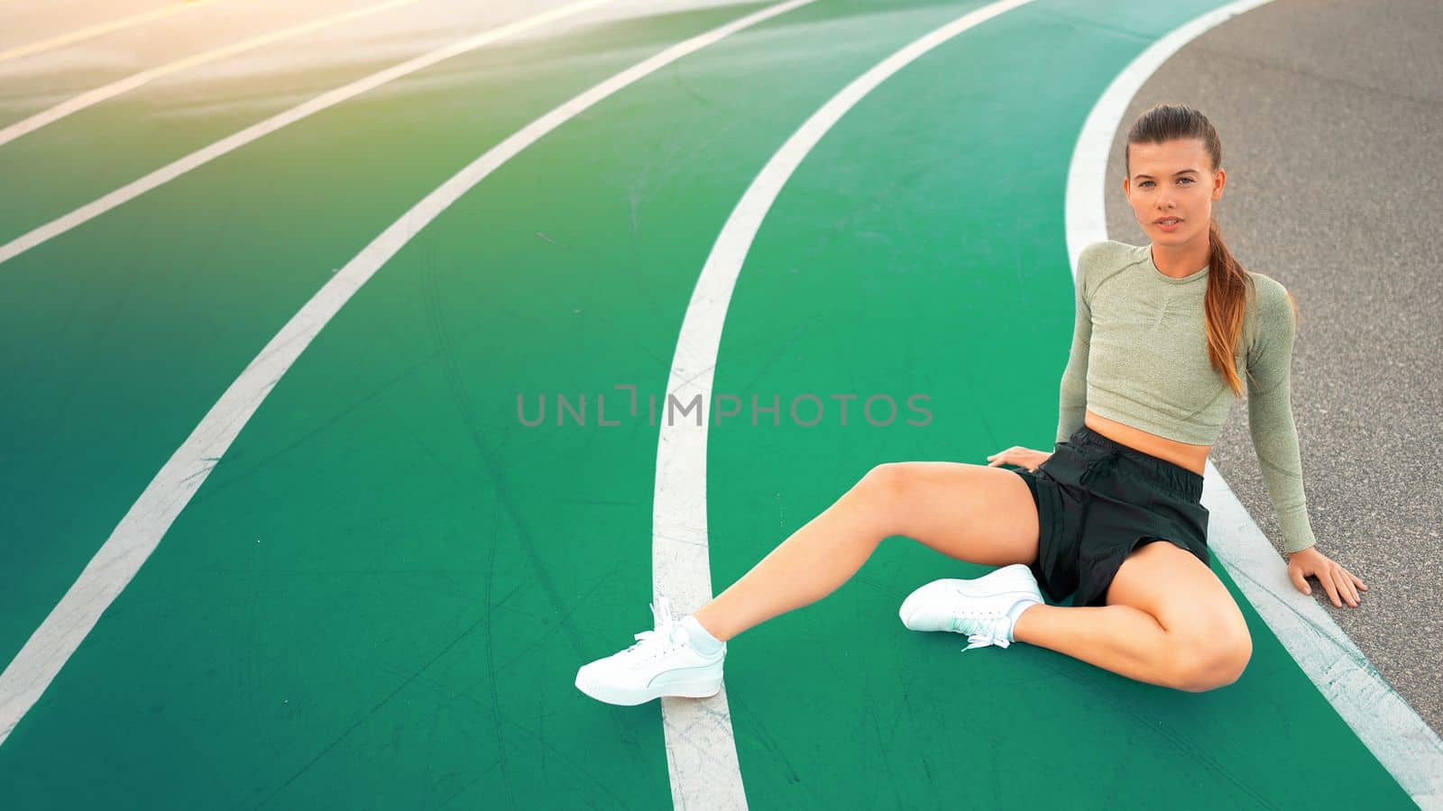 Tired happy woman runner taking rest after run sitting on the running track and smile. Summer evening time. Young athletic female jogger resting after training. Work hard rest harder. Copy space