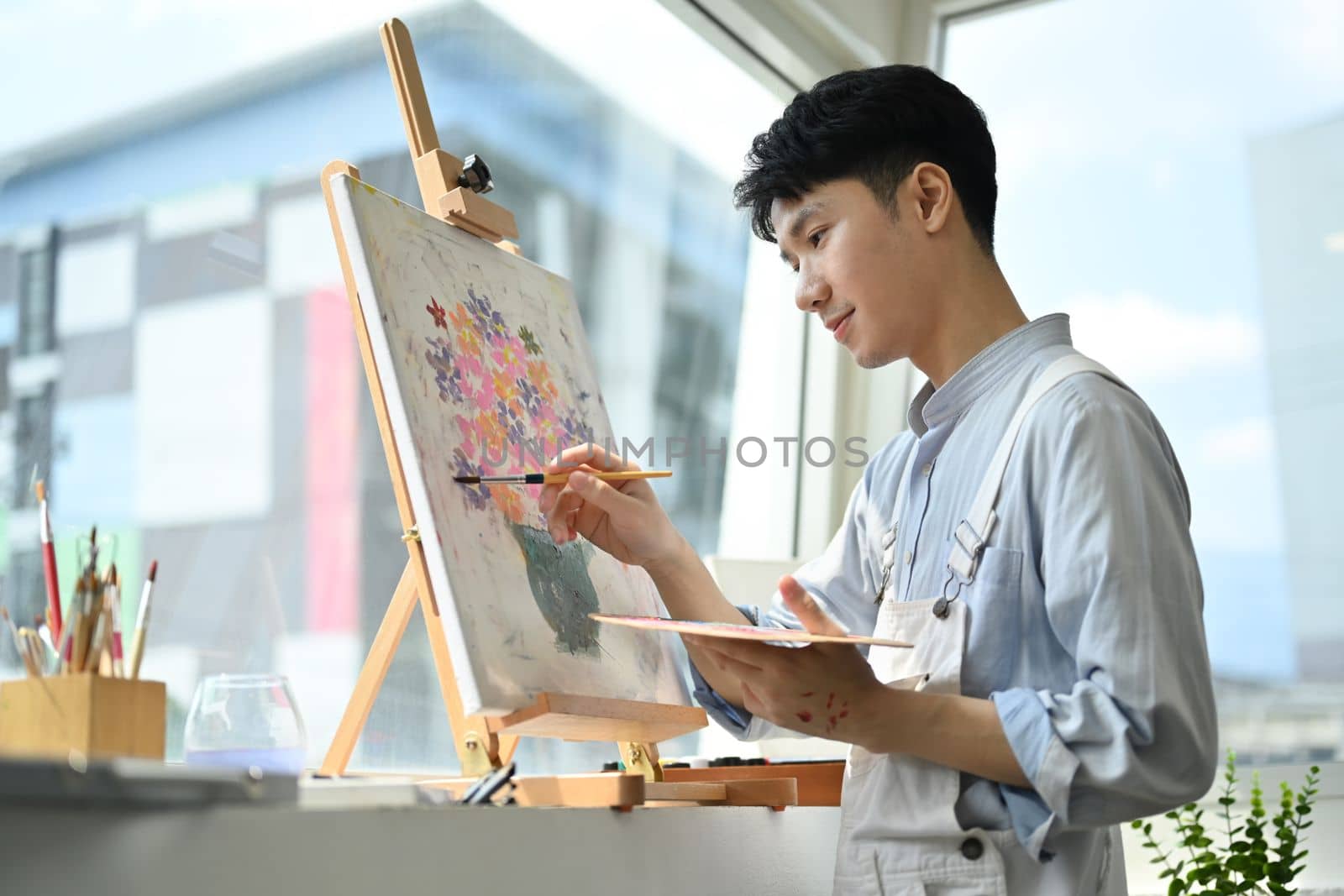 Photo of asian male artist holding paintbrushes and palette painting on easel at bright art studio.