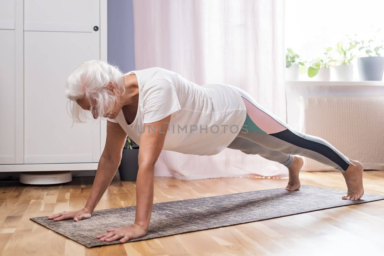 Middle aged woman in plank position keeps fit in her home. Elderly person has fun doing sports
