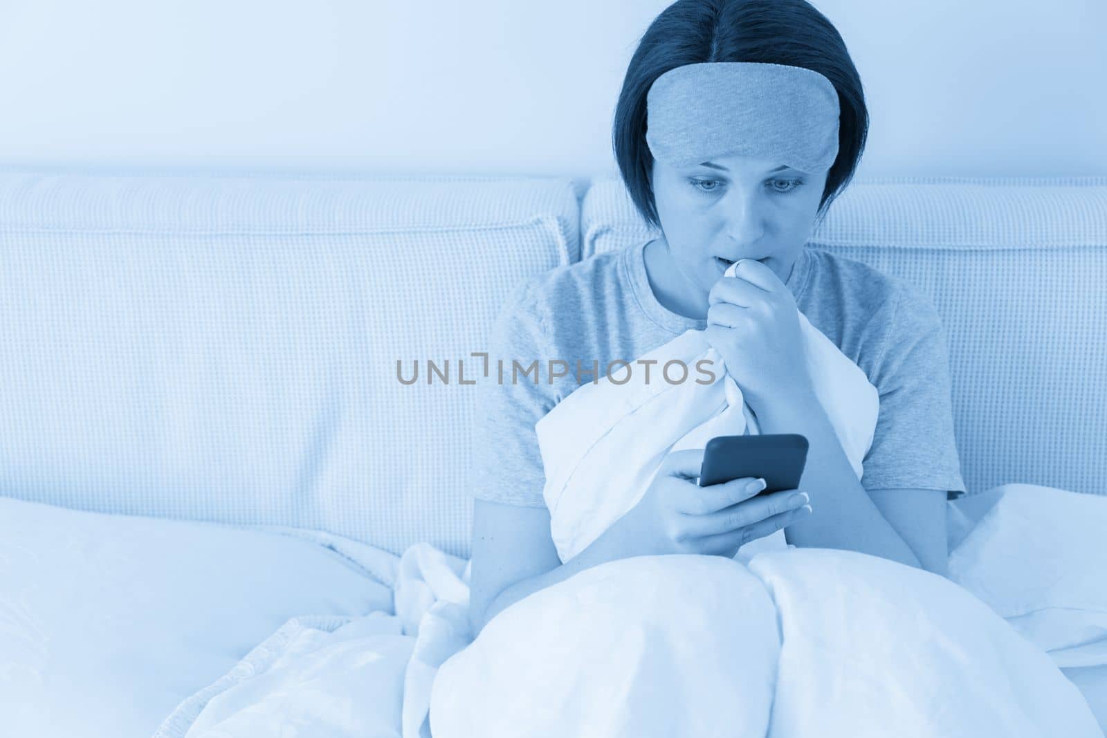Woman wearing sleeping mask texting on smartphone as she sits in bed covered with duvet by Mariakray