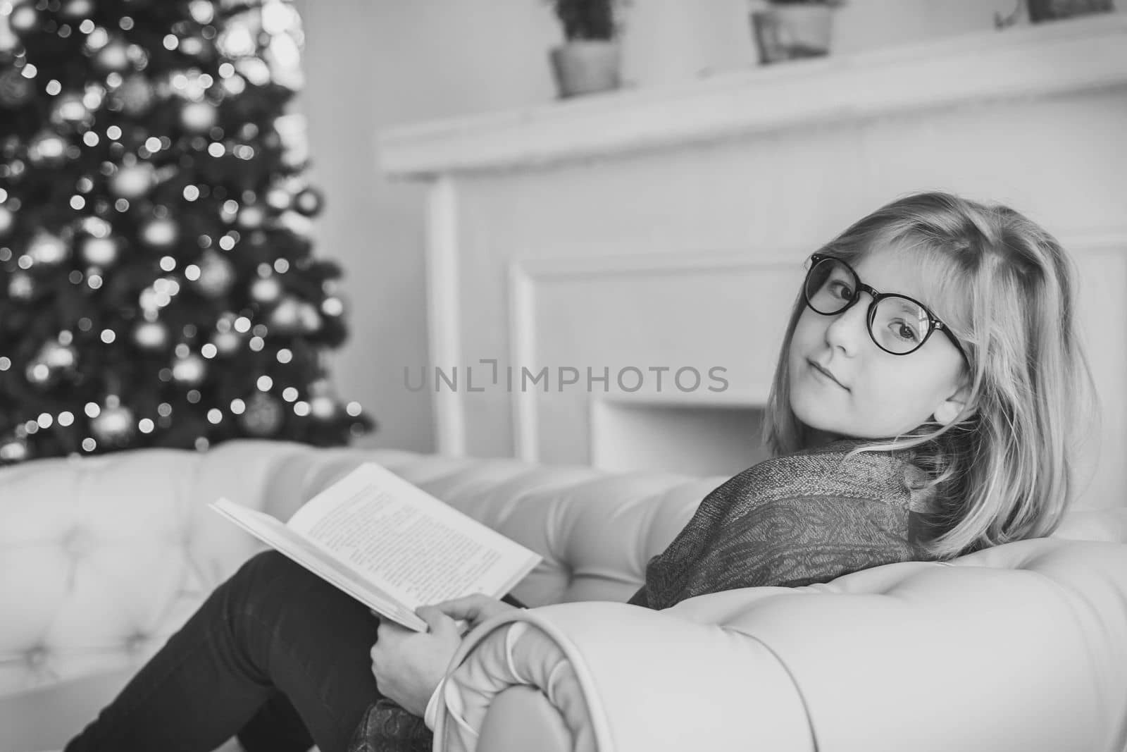 Beautiful girl reading a book on the sofa near the christmas tree.