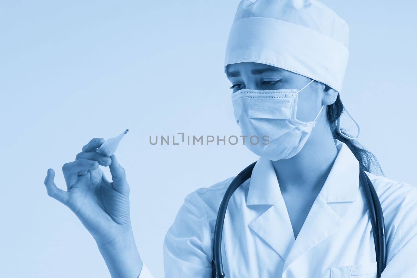 Doctor wearing face mask looking at measurement on medical thermometer over white background by Mariakray