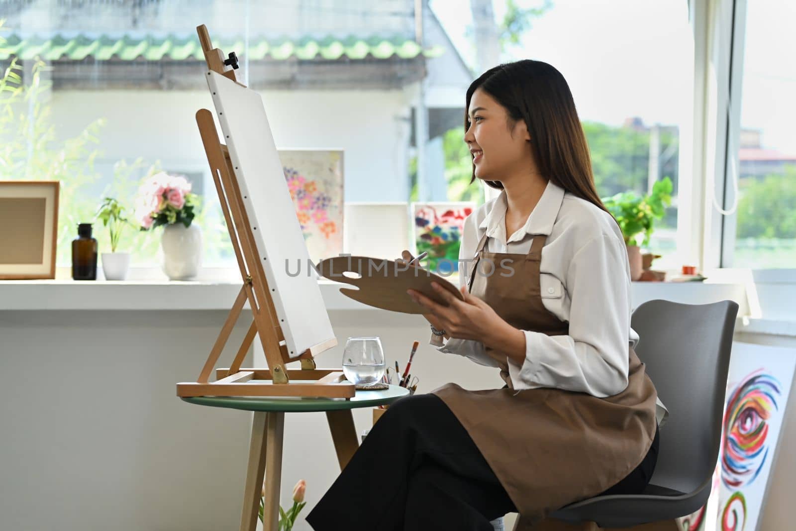 Side view of female painter sitting in front of canvas and painting picture with watercolor. Leisure activity concept.