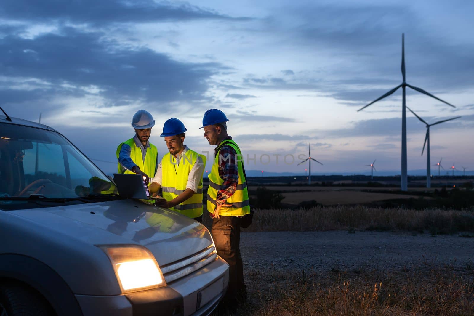 Group of engineers and maintenance workers using laptop together discussing plans in wind turbine farm at sunset. by Hoverstock