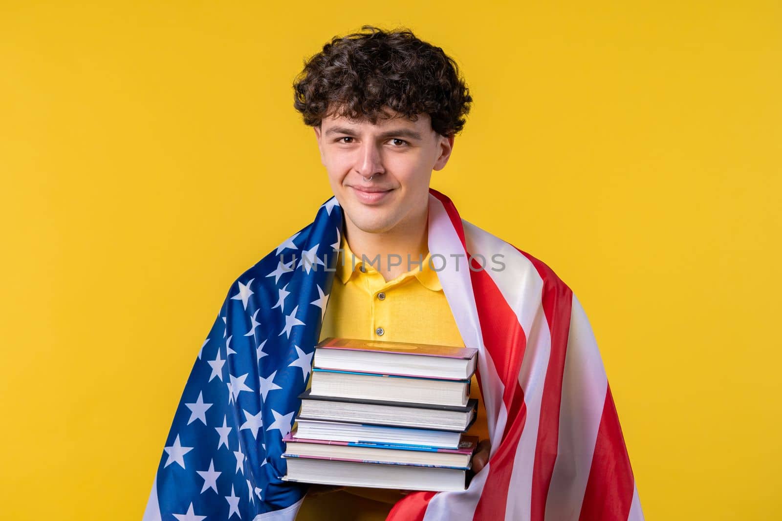 American man student holds stack of university books from college library on yellow background. Happy guy smiles, he is happy to graduate in USA, education abroad concept. High quality photo