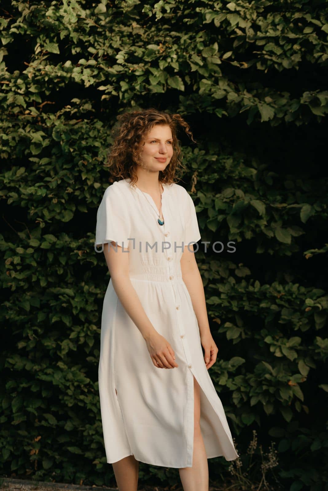 A happy woman with curly hair is posing in the garden in the evening. A lady in a white dress with green leaves in the background.