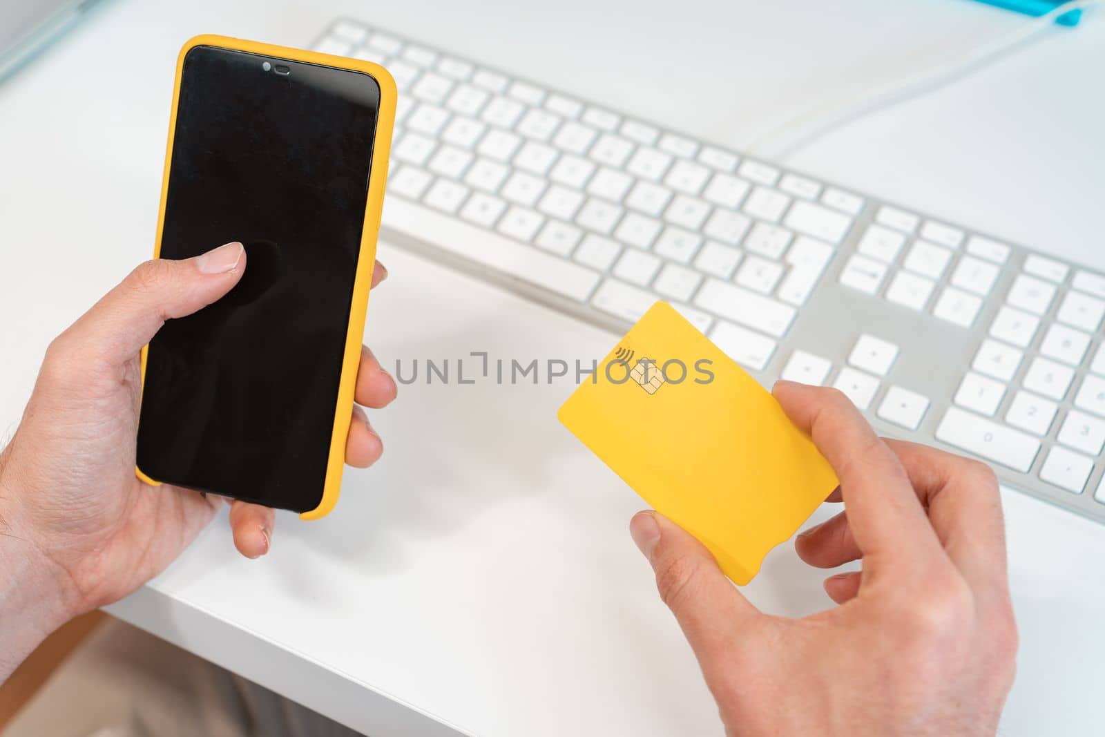 Unrecognizable man paying online with credit card and cell phone in front of a computer at desk office. Yellow and white colors. Smart phone with black screen. High quality photo
