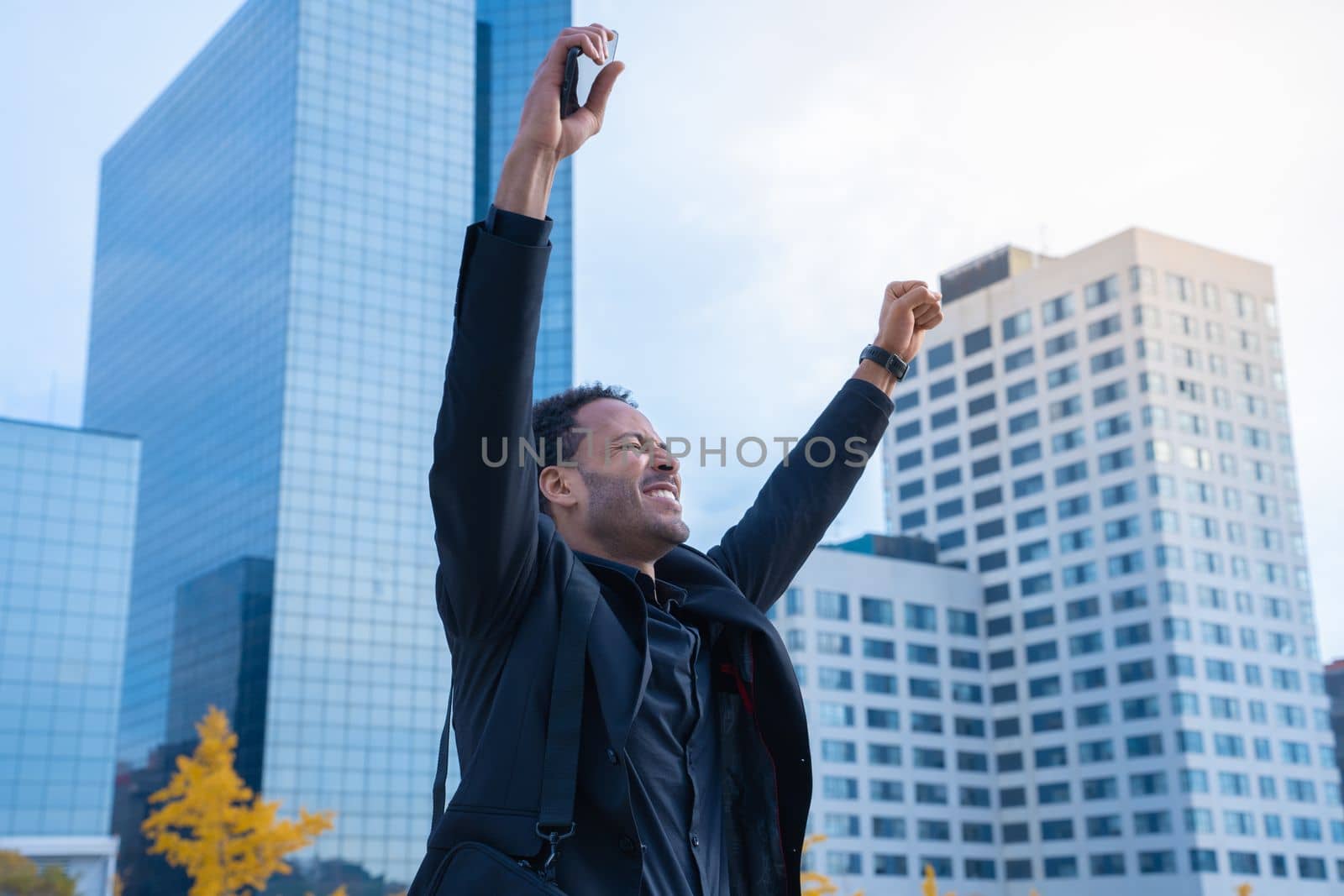 Successful African American businessman in suit raised arms smiling happy celebrates success with financial buildings in background. High quality photo